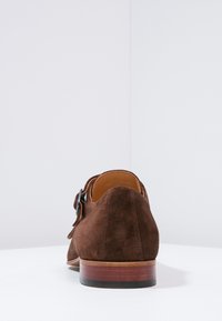 Scarpa in suede marrone con una texture liscia, forma arrotondata e tacco in legno. Presenta una fascetta e un accento in metallo per la chiusura.