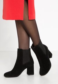 Bottines en suède noir avec un talon bloc, présentant des panneaux latéraux élastiques et une texture lisse. Portées avec des collants noirs et une jupe rouge.