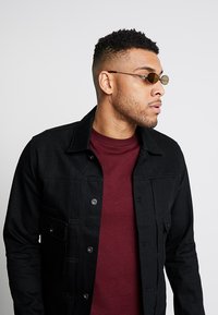 Mustat farkkutakki, jossa on etutaskut ja napit, päällä tumman viininpunainen t-paita; henkilö, jolla on pyöreät, sävytetyt aurinkolasit.