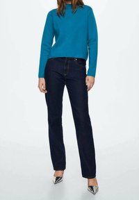 Türkiser Pullover mit langen Ärmeln, kombiniert mit dunkelblauen, schmal geschnittenen Jeans. Das Outfit wird von eleganten silbernen spitzen Pumps ergänzt.