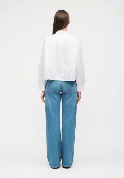 Personne aux cheveux bruns lisses portant un chemisier blanc à manches longues avec une texture au bas du dos et un jean bleu à jambes larges, debout, de dos.