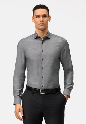 Chemise grise à manches longues avec une texture subtile, devant à boutons et boutons foncés contrastants. Associée à un pantalon noir ajusté et une ceinture noire.