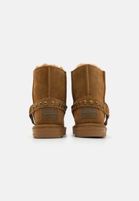 Botas de tobillo de ante marrón con ribete de pelo, tachuelas de metal y un parche de marca en el lateral. Suela de goma plana con un ligero tacón.
