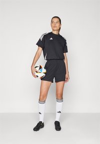 Juodas sportinis marškinėlis ir šortai su baltais dryžiais, juodi sportbačiai ir iki kelių siekiančios baltos kojinės su juodomis juostelėmis. Laiko daugiaspalvį futbolo kamuolį.