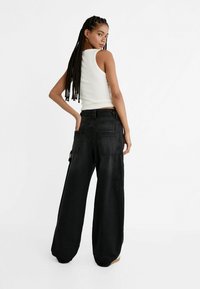 Pantaloni cargo neri a gamba larga con molte tasche, realizzati in tessuto resistente. Il modello indossa un top bianco aderente, con i capelli acconciati in trecce.