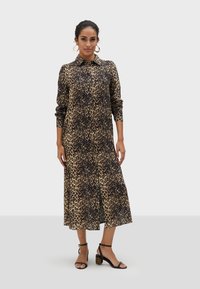 Šaty s leopardím vzorem v béžové a tmavě hnědé barvě, dlouhé rukávy, límeček, volný střih, až k půlce lýtek, doplněné černými sandály na vysokém podpatku.