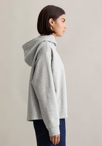 Grijze hoodie met een ontspannen pasvorm, voorzien van een trekkoordcapuchon, geribbelde mouwen en een zachte fleece textuur. Gecombineerd met een donkere spijkerbroek.