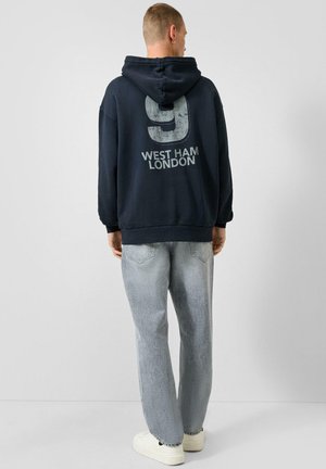 Mann trägt schwarzen Hoodie mit großem Nummer 9 und Text "West Ham London" auf dem Rücken, helle Jeans und weiße Sneakers, steht mit dem Rücken zum Betrachter.