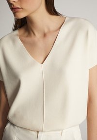 Valkoinen lyhythihainen V-kaula-aukollinen paita, jossa on pehmeä pinta ja hienovarainen sauman yksityiskohta edessä, puettuna valkoisten räätälöityjen alaosien päälle.