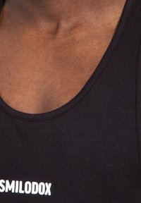 Zwart tanktop met een ronde halslijn, voorzien van witte tekst die "SMILODOX" zegt. De stof lijkt glad en aansluitend.