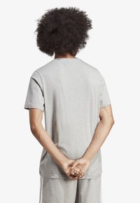 Šedé bavlněné tričko s krátkými rukávy, kulatým výstřihem a volným střihem. Pozadí je jednobarevné bílé. Žádné viditelné vzory ani akcenty.