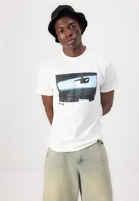 Camiseta blanca de algodón con una impresión gráfica de un interior de coche. Combinada con pantalones cortos de mezclilla claros y un sombrero de cubo negro.