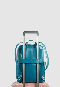 Turquoise leren rugzak met verstelbare riemen en gouden beslag, geplaatst op een roze koffer, met een slank rechthoekig ontwerp en ritssluiting.