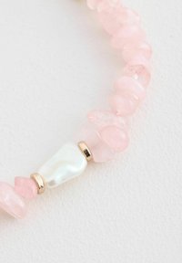 Collar de cuentas con piedras de vidrio rosa, una perla blanca brillante y acentos dorados, dispuestas en un patrón ondulado sobre un fondo blanco texturizado.