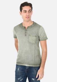 T-shirt verde oliva a maniche corte con scollatura rotonda, tre bottoni e taschino sul petto. Tessuto morbido e vestibilità rilassata. Indossato con jeans blu strappati.