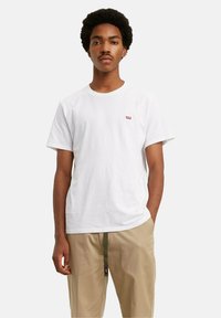 Młody mężczyzna z fryzurą afro, ubrany w białą koszulkę Levi's i beżowe spodnie z oliwkowym paskiem, stojący na jednolitym tle.