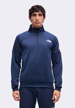 Homme aux cheveux bouclés foncés portant une veste bleu marine à fermeture éclair quart et un pantalon assorti avec les logos The North Face sur la poitrine et la manche.