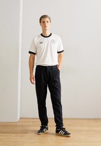 Mand står indendørs iført en hvid ringer t-shirt, sorte bukser og sorte Adidas sneakers med hvide striber på trægulv.