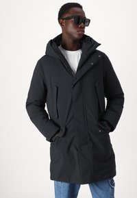 Parka nera isolata con zip frontale, cappuccio ampio e tasche laterali. Indossata sopra una camicia bianca con dettagli di cucitura visibili.