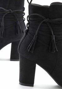 Stivaletti neri in suede alla caviglia con tacco a blocco alto, caratterizzati da dettagli laterali arricciati e nappine legate sul retro. Texture liscia, punta affusolata.