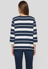 Frau mit glattem braunem Haar, die ein navyblau und weiß gestreiftes langärmliges Shirt und dunkle blaue Hosen trägt, steht mit dem Rücken zugewandt.