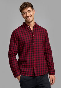 Camisa de franela a cuadros rojos y negros con collar botón, mangas largas y detalles de botones de madera. Se lleva con pantalones oscuros.