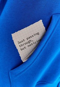 Niebieska bawełniana bluza z kapturem z przednią kieszenią. Widoczna jest kartka z postrzępionym brzegiem, na której znajduje się czarny tekst na tle w kratkę.