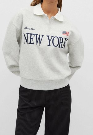 Osoba ubrana w jasnoszarą bluzę z białym kołnierzem, z napisem "NEW YORK", małą flagą USA oraz wyszywanym napisem "Manhattan", w połączeniu z czarnymi spodniami w cienkie prążki.