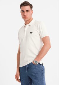 Vit kortärmad polo med en svart triangulär logotyp på vänster bröst, i kombination med blå jeans. Slät tygtextur.