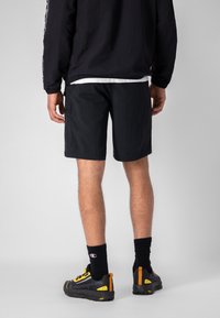 Pantaloni corti neri con una vestibilità comoda, realizzati in materiale leggero. Indossati con calze nere e scarpe con dettagli gialli e branding.