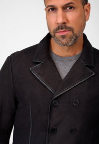 Manteau en cuir noir à double boutonnage, avec un large col et des détails de couture visibles. Pull gris porté en dessous.