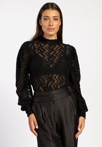 Schwarzes Spitze-Top mit Zickzackmuster, hohem Kragen und puffigen langen Ärmeln, kombiniert mit dunklen, satinartigen, hoch taillierten Hosen.