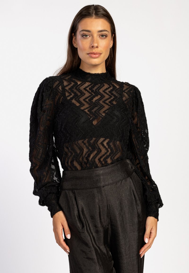 Schwarzes Spitze-Top mit Zickzackmuster, hohem Kragen und puffigen langen Ärmeln, kombiniert mit dunklen, satinartigen, hoch taillierten Hosen.