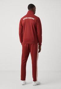 Crvena trenirka s visokim ovratnikom; "BAYERN MUNCHEN" ispisano na leđima. Ima bijele pruge niz hlače. Izrađena je od glatkog materijala.