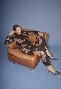 Vrouw leunt achterover op een bruin leren fauteuil, gekleed in een zwarte bloemenoutfit en zilveren puntige hakken, tegen een blauwe achtergrond.