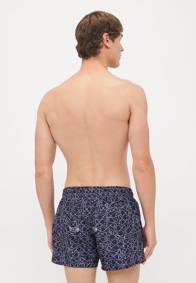 Ein oberkörperfreier Mann, der mit dem Rücken zur Kamera steht, trägt marineblaue Badehosen mit weißem Fischmuster und zwei Gesäßtaschen vor einem schlichten weißen Hintergrund.