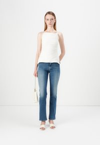 Witte, aansluitende tanktop met een vierkante halslijn en gerimpelde details, gecombineerd met lichtblauwe rechte jeans en beige instapsandalen.