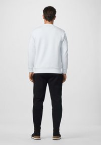 Uomo in piedi, di spalle, che indossa una felpa azzurra chiara, pantaloni neri aderenti e sneakers nere con suola marrone, su uno sfondo neutro.