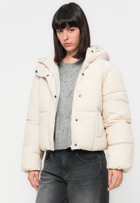 Krem pufferjakke med hette, med glidelås og trykknapper, quiltet design og to frontlommer. Teksturert, mykt stoff.
