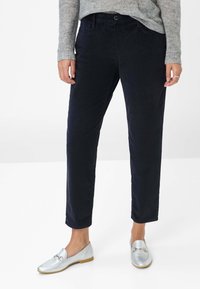 Femme portant un pantalon en velours côtelé bleu marine, un pull gris clair à motifs et des mocassins argentés, se tenant sur un sol blanc.