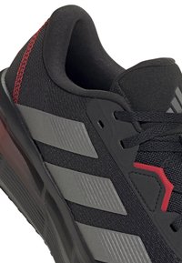 Chaussure de sport noire avec des bandes diagonales grises, coutures rouges au talon, col rembourré et lacets noirs sur une surface supérieure texturée.
