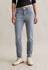 Vrouw in lichtgrijze slim-fit jeans en beige sneakers, staande tegen een effen neutrale achtergrond.