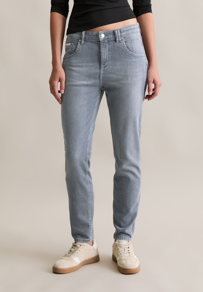 Vrouw in lichtgrijze slim-fit jeans en beige sneakers, staande tegen een effen neutrale achtergrond.