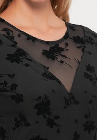 Schwarzes Oberteil mit durchscheinendem Mesh und schwarzem Blumenmuster, runder Ausschnitt und V-förmigem Ausschnitt. Weicher Stoff mit bestickten Details.