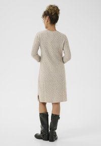Vrouw met een krullende bun, gekleed in een beige textuur gebreide jurk en zwarte leren laarzen tot halverwege het onderbeen, staande met de rug naar de camera tegen een effen achtergrond.