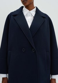 Marineblauer doppelt gefütterter Mantel aus weichem Stoff, mit umgeschlagenem Kragen, zwei vorderen Taschen und einem lässigen Schnitt.