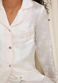Top in satin bianco con un delicato motivo a fiocchi, featuring piping rosa pallido, una tasca sul petto e bottoni argentati lungo la parte anteriore.