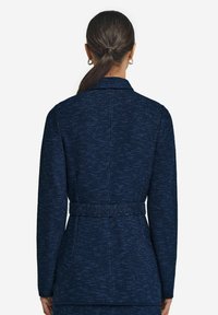 Marinblå, texturerad kavaj med knytbälte i midjan, långa ärmar och slagkrage. Bakifrån framhävs tygets intrikata detaljer.