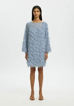 Mujer de pie, vestida con un vestido azul claro hasta la rodilla con detalles texturizados similares a plumas y mangas largas, combinado con sandalias beige de punta abierta.