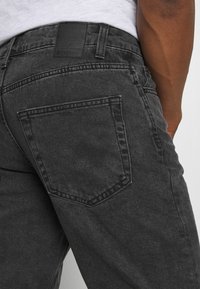 Jean en denim charbon avec une poche arrière ornée de coutures et une étiquette en cuir noir au niveau de la taille. Fini en tissu texturé.